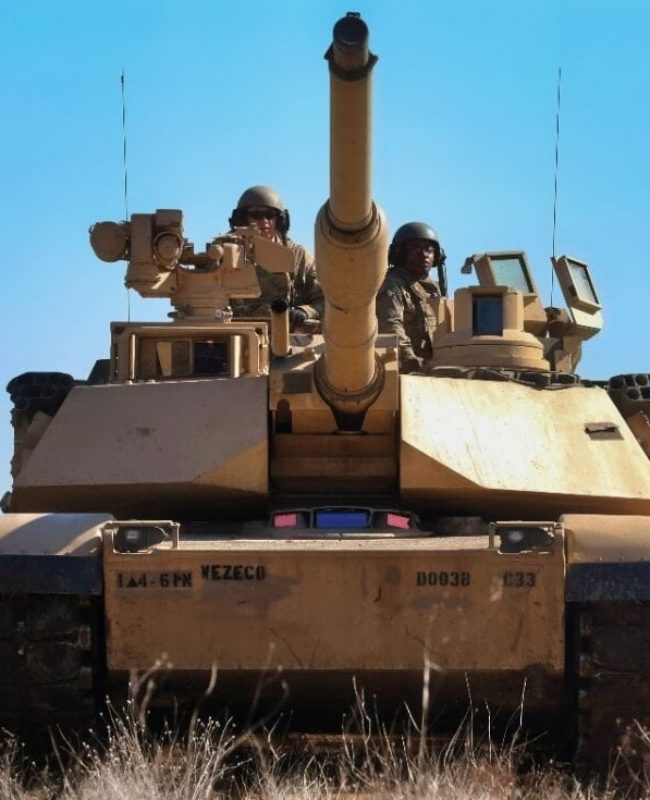 A tan armored tank with a long gun barrel pointing forward, crew in helmets on the turret, in a dry, open field with a line of soldiers nearby.